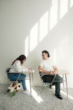 a man and woman sitting at desks in front of a white wall with sunlight coming through the windows