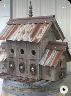 a wooden birdhouse sitting on top of a barrel next to a white building with rusted metal roof