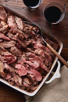 a roasting pan filled with meat next to two glasses of wine on top of a wooden table