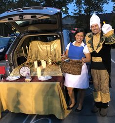 two people in costumes standing next to a car with its trunk open and food on the table