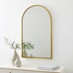 a white dresser topped with a mirror and vase filled with flowers next to a plant