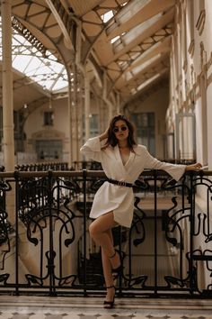 a woman leaning on a railing with her arms behind her head, wearing sunglasses and a white dress