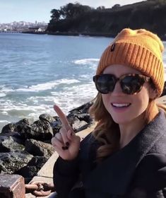 a woman wearing sunglasses and a hat is making the peace sign with her fingers by the ocean