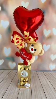 a teddy bear holding a heart shaped balloon in it's mouth and sitting on top of a box