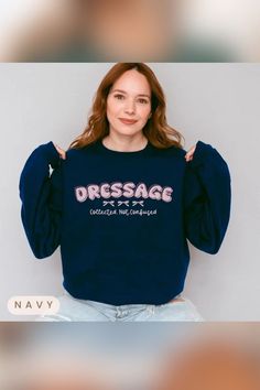 A woman with long, wavy red hair is wearing a navy blue sweatshirt that reads "DRESSAGE" in large pink letters with three bows underneath. Below the bows, the text says, "Collected. Not Confused." She is sitting casually in ripped light-wash jeans and lightly smiling at the camera. The word "NAVY" is displayed in a small beige label in the bottom left corner. The Stables, A Horse