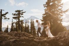 a bride and groom holding hands on top of a mountain with trees in the background