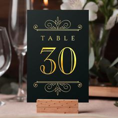 a table number sign sitting on top of a wooden block next to wine glasses and flowers