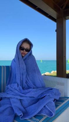 a woman wrapped in a blue towel sitting on a bench next to the ocean and palm trees