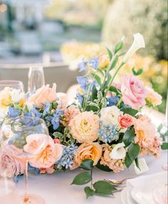 an arrangement of flowers and greenery on a table with wine glasses in the foreground