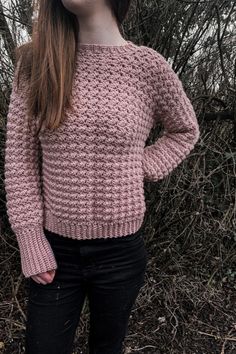 a woman standing in front of some bushes wearing a pink knitted sweater and black jeans