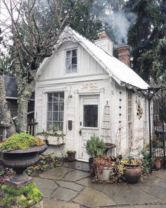 a small white house sitting in the middle of a yard with lots of potted plants