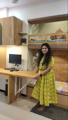 a woman standing in front of a desk with a laptop on it and a bed behind her