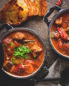 two black pots filled with stew next to bread and parsley on a wooden table