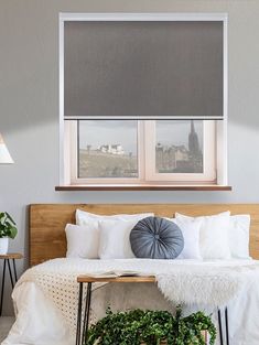 a bed with white sheets, pillows and plants in front of two windows that have roman shades on them