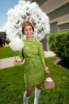 a woman in green dress and white feather hat holding an umbrella over her head while standing on grass