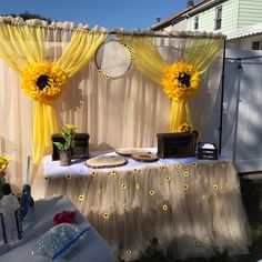 a table with yellow curtains and sunflowers on it in front of a house