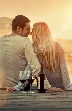 a man and woman sitting on a dock with wine glasses in front of their faces