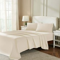 a bed with white sheets and pillows next to a lamp on a table in front of a window