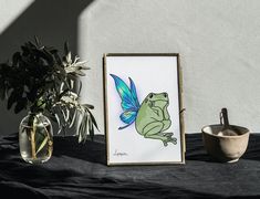 a frog is sitting next to a potted plant and a framed art print on a table