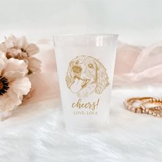 a glass with a dog's face on it next to flowers and a ring