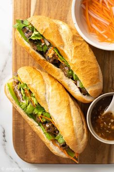 two sub sandwiches sitting on top of a cutting board next to a bowl of cole slaw