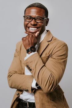 a man in a tan jacket and glasses smiles while leaning his chin on his hand