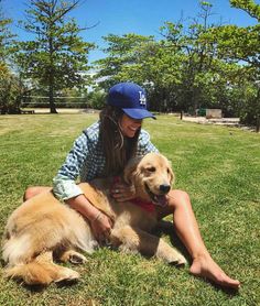 a woman sitting in the grass petting a brown dog with a blue hat on