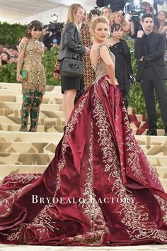 a woman in a red and gold gown standing on steps with people taking pictures behind her