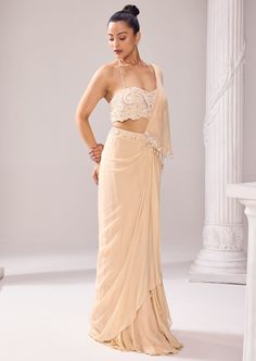 A drape saree in muted beige hues allured with intricate embroidery on a tube blouse with a halter neck beaded strap. There is a cascade drape attached from the belt, falling free from the front. The distinctive artwork with ethereal detailing in the blouse and hemline of the cape exhibits impeccable couture craftsmanship. The ensemble is further highlighted with tassels all over the pallu and cascade. Elegant Side Open Georgette Choli, Elegant Beige Choli With Traditional Drape, Traditional Drape Beige Georgette Lehenga, Beige Georgette Lehenga, Beige Lehenga With Traditional Drape In Georgette, Beige Lehenga In Traditional Drape Style, Designer Pre-draped Cream Saree, Designer Cream Pre-draped Saree In Traditional Style, Designer Cream Pre-draped Saree