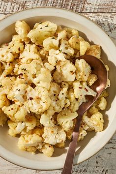 a white bowl filled with cauliflower on top of a wooden table next to a spoon