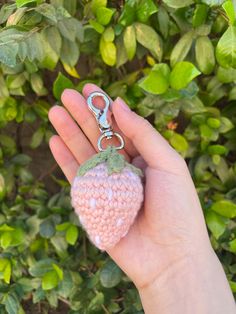 a hand holding a tiny crocheted strawberry keychain in front of green leaves