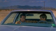 two men sitting in the driver's seat of a car on a desert road