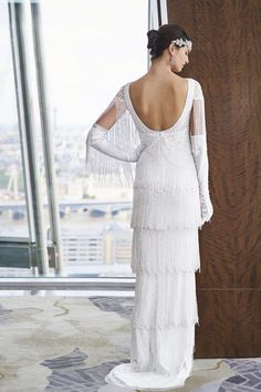 a woman in a white dress standing next to a tall window looking out at the city