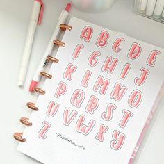 an open notebook with pink letters on it next to some pens and other items that include markers