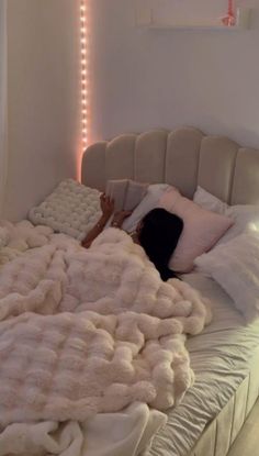 a woman laying on top of a bed covered in blankets and pillows with lights behind her