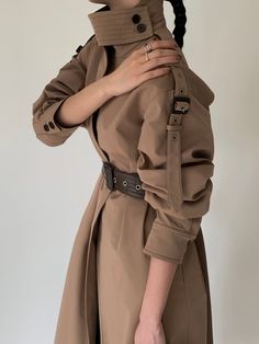 a woman wearing a trench coat and holding her hand on her shoulder with both hands