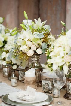 the table is set with silver vases and white flowers in them, along with other place settings