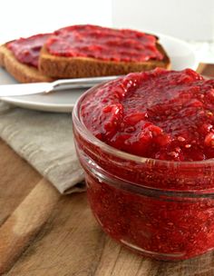 a jar of strawberry jam sitting on top of a wooden table next to a slice of bread