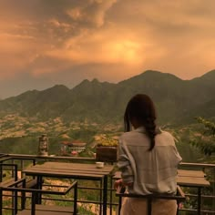 a woman standing on top of a balcony next to a lush green hillside under a cloudy sky