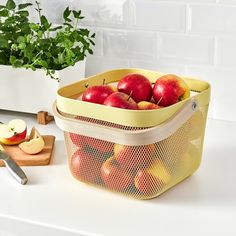 a yellow mesh basket filled with apples next to a cutting board and potted plant