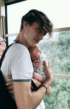 a man holding a baby in his arms looking out a window at the trees outside