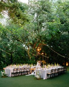 an outdoor dinner table set up with white linens and lights strung from the trees