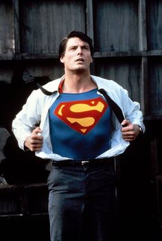 the man is wearing a superman t - shirt with his hands on his hips while standing in front of a wooden wall