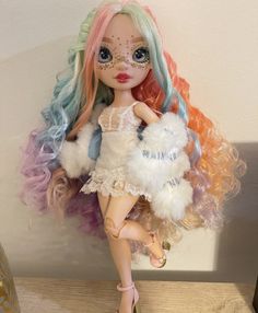 a doll with multicolored hair is holding a stuffed animal in her hand and sitting on a table