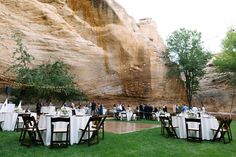 a group of people sitting at tables in front of a large rock wall with lights strung from it