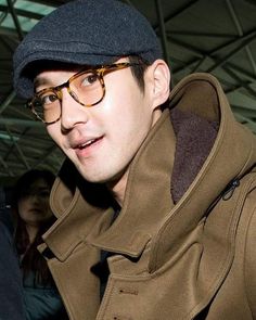 a man wearing glasses and a hat with a coat over his shoulders is looking at the camera