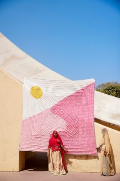 two women standing in front of a large pink and white quilted wall hanging on the side of a building