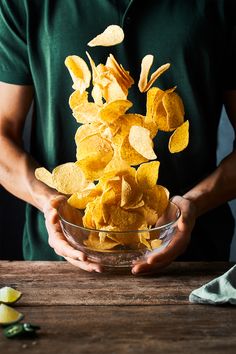 a man holding a bowl full of potato chips on top of a wooden table next to sliced lemons