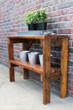 a wooden table with cups on it next to a brick wall and potted plant