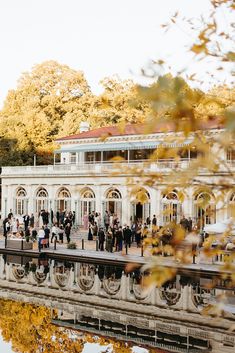 many people are standing on the side of a building near water and trees with yellow leaves